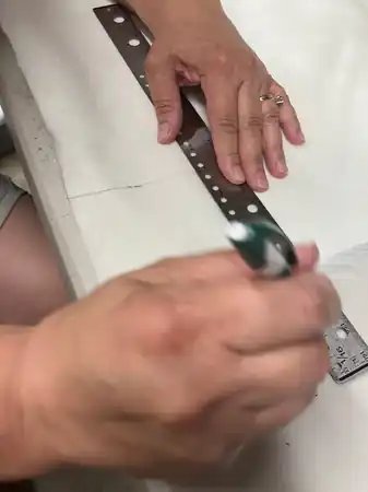 Hands marking a dotted pattern line on purple floral fabric with a pencil, pins and measuring tape on the table