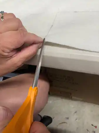 Hands using orange-handled scissors to cut cream fabric along a marked line at the work table